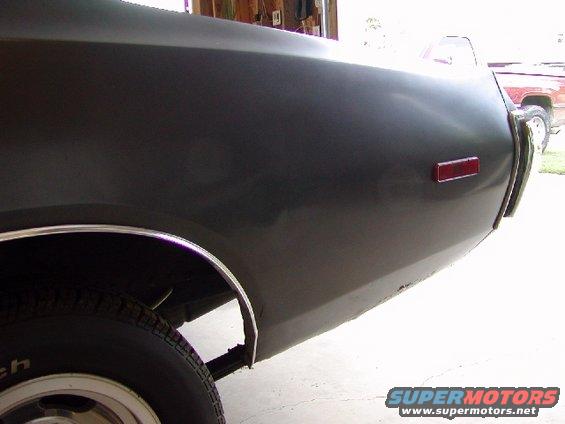 Dark green car's rear quarter panel and taillight, showing rust near the bottom edge. A red pickup truck is visible in the background