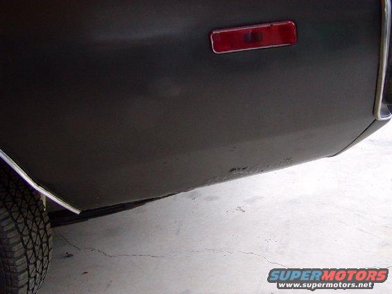 Close-up of dark-colored rear quarter panel with a red reflector and visible rust spots near the bottom edge