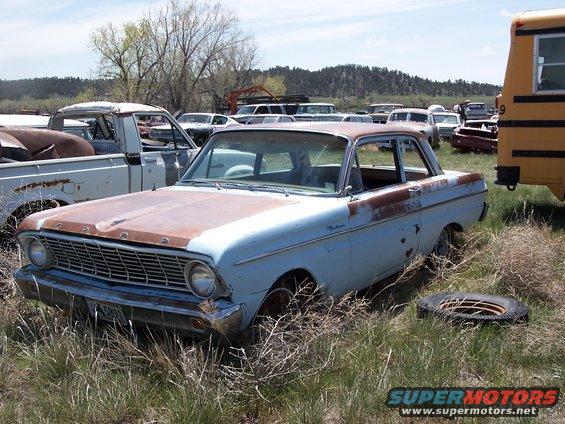 Light blue sedan with rusty hood and roof, seen from a 3/4 front angle in a junkyard