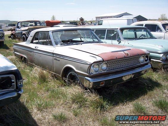 Silver coupe with a rusty hood and cracked windshield sits in a junkyard