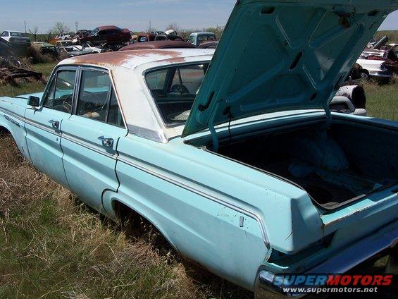 Light blue sedan with open trunk, sitting in a junkyard with other cars visible