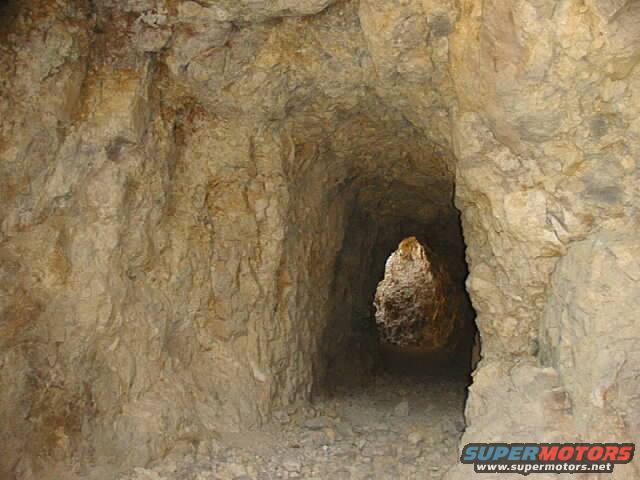 Mine tunnel with rough rock walls and a bright opening in the distance. Supermotors.net logo in the corner
