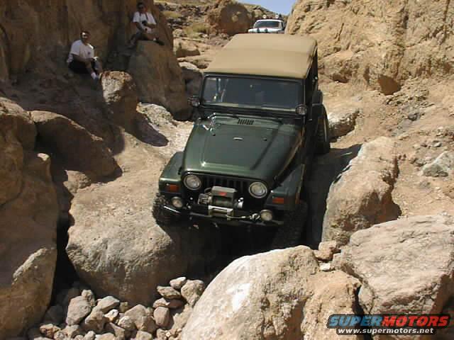 Green Jeep Wrangler navigating a rocky trail with two people sitting on rocks nearby