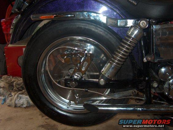 Motorcycle rear wheel with chrome rim, disc brake, and coil-over shock absorber. Purple fender above