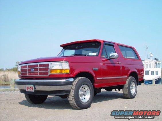 Red 1995 Ford Bronco parked by a body of water with a boat in the background