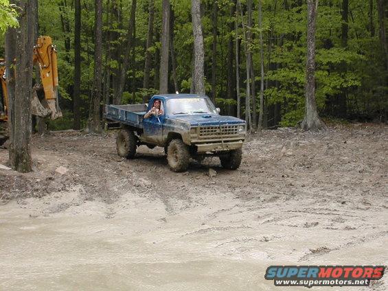 Blue pickup truck with a flatbed driving through a muddy wooded area. Driver is visible