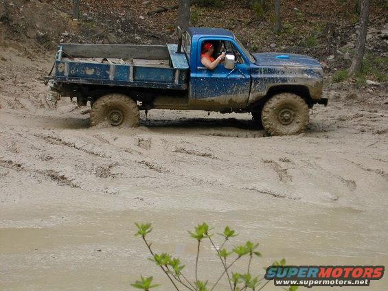 Blue pickup truck with a flatbed is stuck in thick mud. Driver wearing a red bandana is behind the wheel