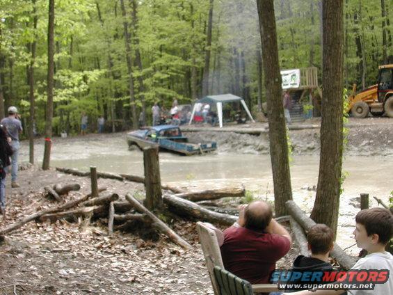 Blue pickup truck stuck in mud, side view, with people watching and a backhoe in the background