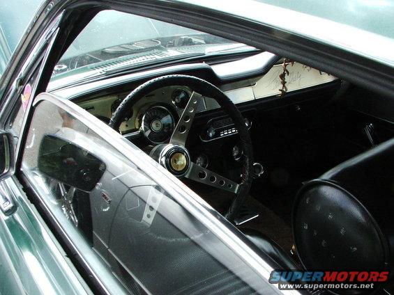 Interior view of a classic car, showing the steering wheel, dashboard, and seats. The steering wheel has a gold emblem