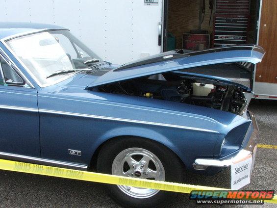 Blue classic car, hood open, showing engine bay. GT badge visible on fender. Yellow caution tape in foreground