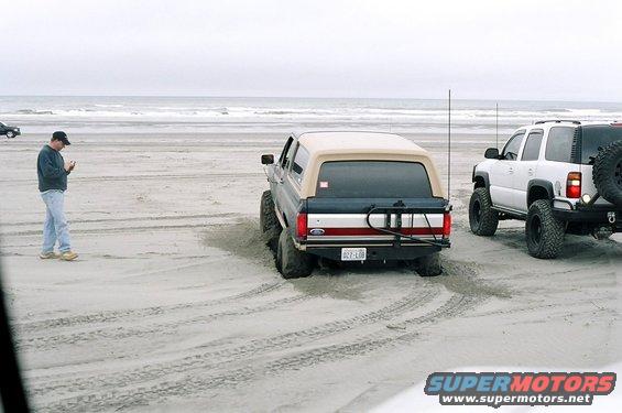 Gray Ford Bronco with tan top stuck in sand, rear view. Red stripe on tailgate