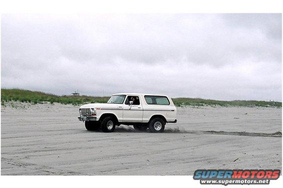 White Ford Bronco driving on a sandy beach with dunes and overcast sky