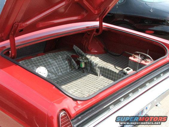 Red car trunk open, showing plaid-lined interior with spare tire and battery. Black car visible in background