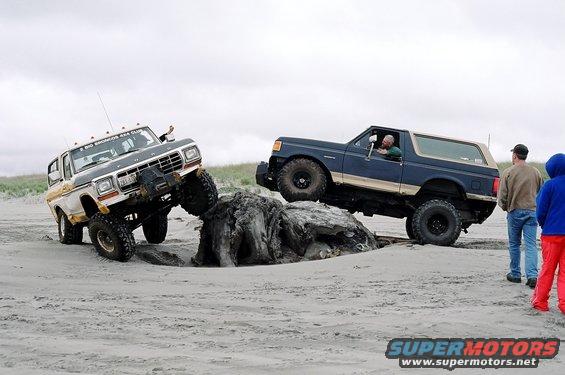 Two 1979 Ford Broncos off-roading on a beach, one climbing over driftwood