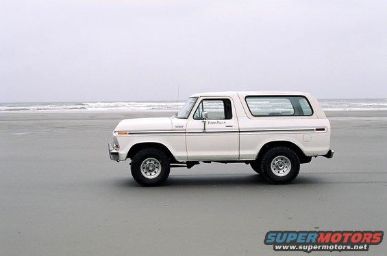 White Ford Bronco parked on a sandy beach with the ocean in the background. "Ford" is visible on the door