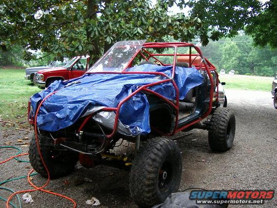 Buggy with red roll cage and blue tarp on front, parked outside with other vehicles
