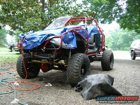 Off-road vehicle with blue tarp and red roll cage, large tires, parked on gravel