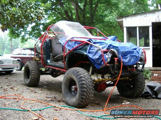 Off-road vehicle with roll cage, tarp, and large tires. Another vehicle visible