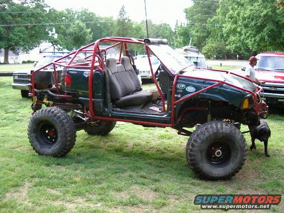 Custom off-road buggy with red roll cage, dark green body, and large tires. Another truck is visible behind it