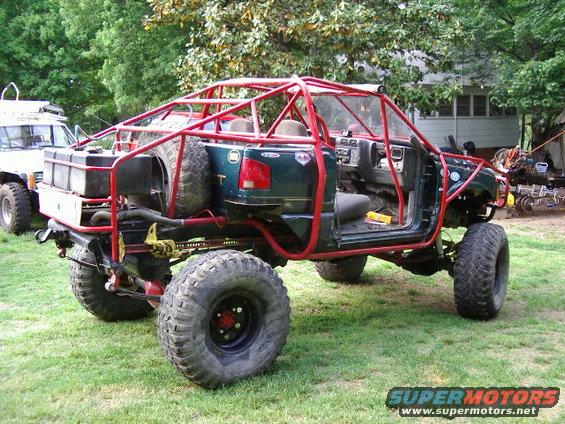 Heavily modified green truck with a red roll cage and large off-road tires. Another white SUV is parked behind it