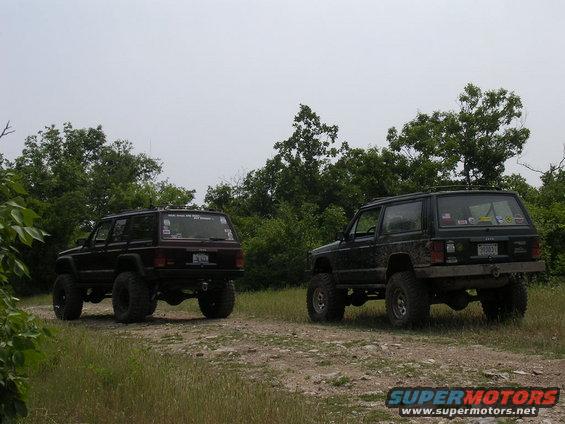 Two lifted Jeep Cherokees parked on a dirt path with trees in the background. One is dark red, the other is dark green