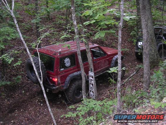 Red 1995 Jeep Cherokee with spare tire on back, driving on a wooded trail. Another dark SUV is visible behind it