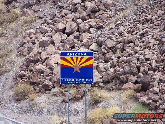 Arizona state sign with flag and text, surrounded by rocks and dry brush