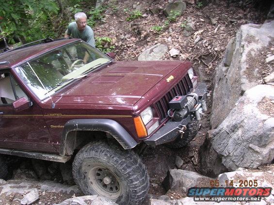 Maroon Jeep Cherokee with winch navigating a rocky trail. Man in green shirt watches