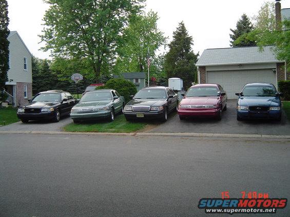 Five Ford Crown Victoria sedans parked in a driveway, varying colors including black, green, maroon, and blue. The front of each vehicle is visible