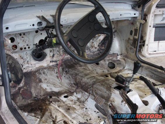 Car interior stripped down, showing bare metal floor, steering wheel, and gear shift. Appears to be from the late 80s or early 90s