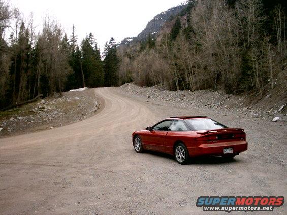 Red 1996 Ford Probe parked on a winding dirt road with trees and mountains in the background