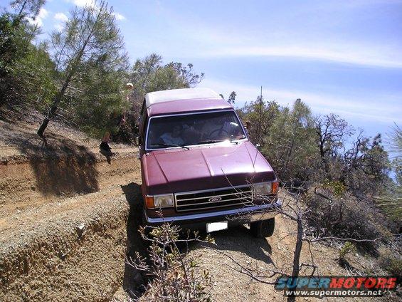 Maroon Ford Bronco driving on a dirt trail with trees and bushes. A person is visible in the driver's seat