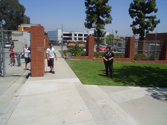 School entrance with a chain-link fence, brick pillars, and a police officer standing on the grass