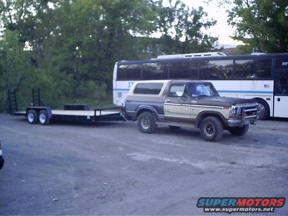 1979 Ford Bronco with two-tone paint, towing a flatbed trailer. Bus in background