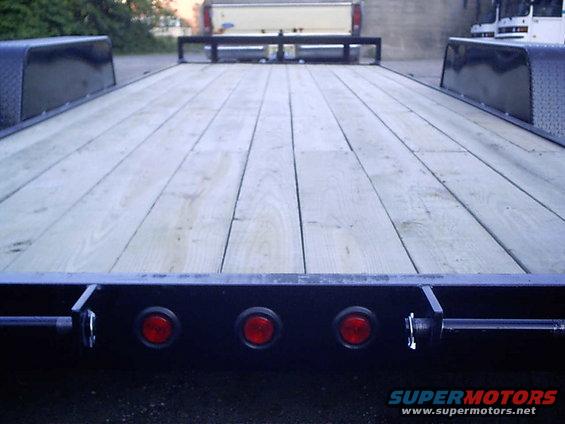 Flatbed trailer with wooden deck, three red lights on rear bumper. Pickup truck visible in background
