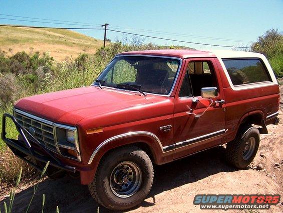 Red Ford Bronco SUV with white top, front-left view on dirt trail