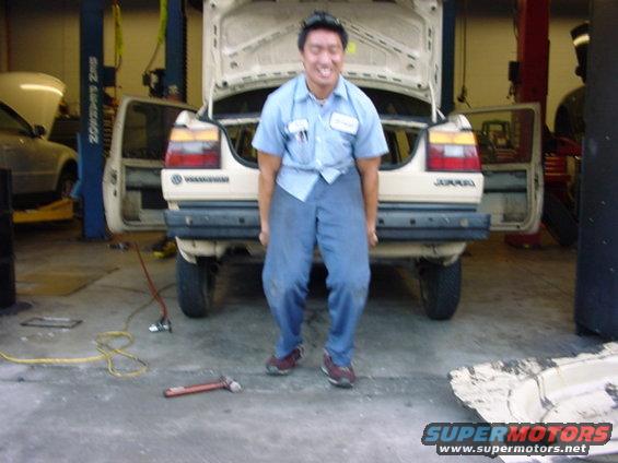 Man in blue shirt and pants lifting beige Volkswagen Jetta's rear bumper. The car's trunk is open