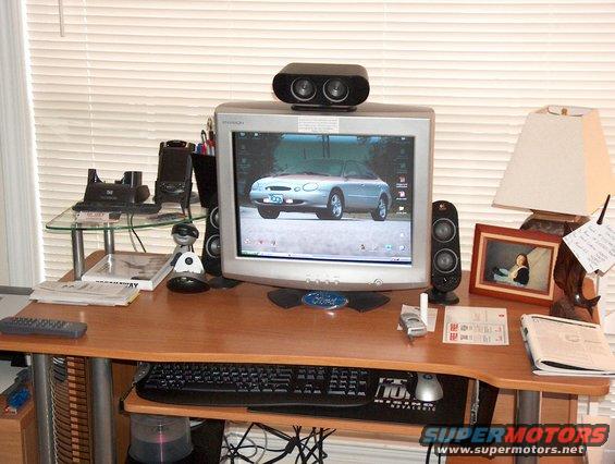 Desktop computer displaying a silver car on its screen, with speakers, keyboard, and other office items