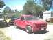1987 Ford Bronco - A red pickup truck tows a yellow car on a trailer, with a trailer home and trees in the background.