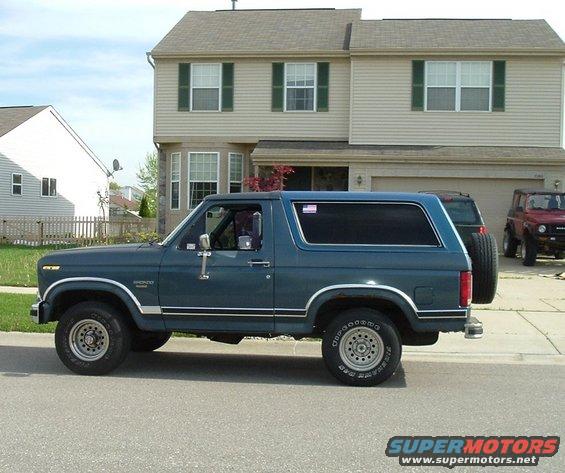 Dark blue Ford Bronco parked on street, house in background. Another SUV and a red Jeep visible in driveway