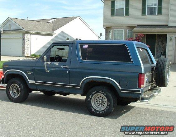 Blue 1980s Ford Bronco SUV parked on street, side view, spare tire on rear door