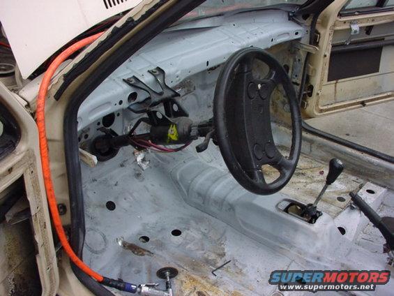 Car interior stripped down, showing bare metal floor, steering wheel, and shifter. Orange hose and air tool visible