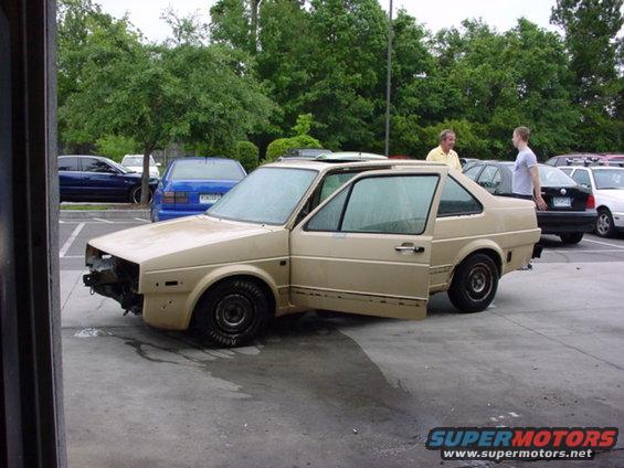 Tan Volkswagen sedan, doors open, missing front bumper, sunroof, parked in lot