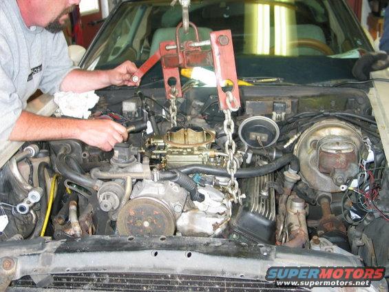 Engine bay with engine hoist attached to the engine, mechanic working on carburetor