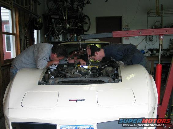 Two guys leaning over the engine bay of a white 1981 Chevrolet Corvette, working on it with an engine hoist overhead