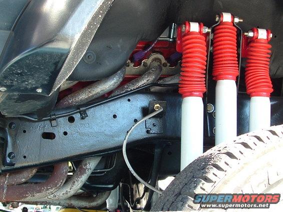 Underside of a vehicle showing the frame, exhaust pipes, and three red and white shock absorbers. A tire is visible on the right