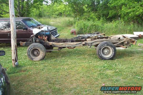 Chassis of a vehicle with engine and front wheels, body removed. Another vehicle is partially visible on the left