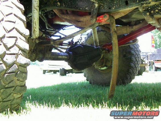 Underside of a lifted truck with large tires, showing suspension and drivetrain components. Another vehicle is visible in the background