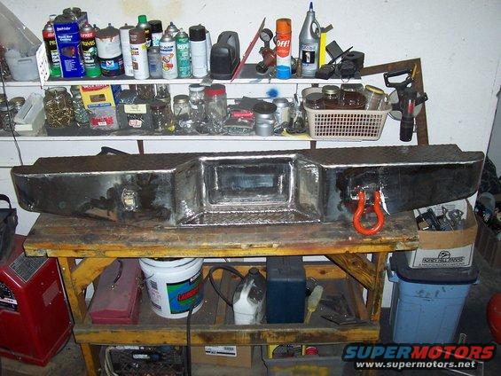 Aftermarket chrome truck bumper with a red tow hook attached, resting on a workbench