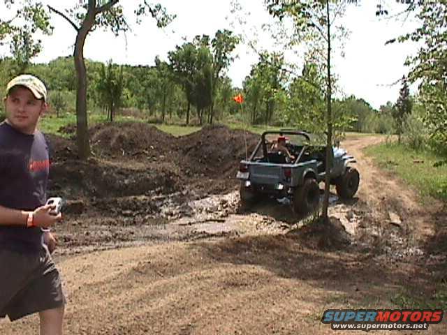 Guy in a baseball cap watches a Jeep Wrangler drive through mud on a dirt path